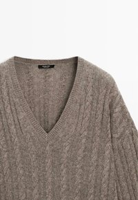 Maglione a V in una tonalità di marrone tenue, caratterizzato da un design a trecce e finitura texture. Tessuto morbido con bordi a coste.
