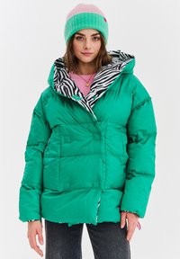 Žena v zelenej bunda s čiernym a bielym vzorom zebry vo vnútri, ružovom svetri a zelenej a ružovej čiapke, stojí na bielom pozadí.