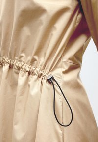 Tissu beige avec taille élastique froncée resserrée par un cordon noir réglable et un embout argenté sur le côté droit du vêtement.