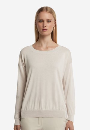 Frau mit glatten blonden Haaren, die goldene Creolen und einen lockeren, hellbeigen Langarmpullover mit rundem Ausschnitt trägt.