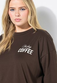 Brun bomulds T-shirt med korte ærmer, med hvid tekst der siger "Fredagskaffe" i et afslappet skrifttype. Glat tekstur, løs pasform.