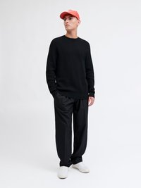 Ung man som bär en texturerad svart tröja, svarta byxor, vita sneakers och en röd keps, stående mot en enkel ljus bakgrund.