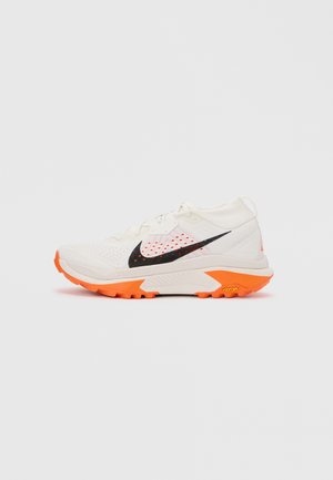 Άσπρο και πορτοκαλί παπούτσι τρεξίματος Nike με μαύρο λογότυπο swoosh και υφή στη σόλα, σε απλό γκρι φόντο.