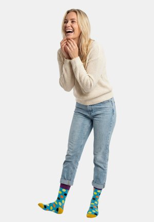 Blondynka śmiejąca się, ubrana w kremowy sweter z dzianiny, jasnoniebieskie podwinięte dżinsy oraz kolorowe skarpetki w kropki, stojąca na białym tle.