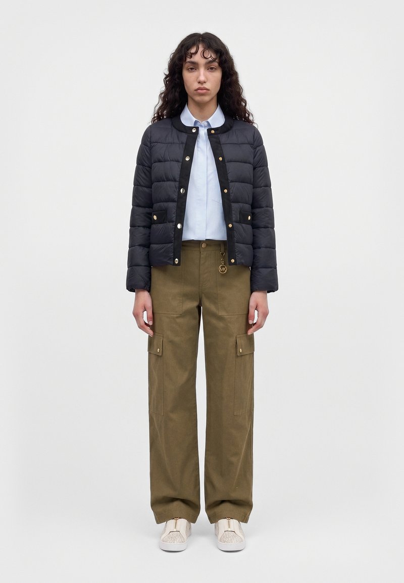 Un mannequin se tient devant un fond uni, vêtu d’une veste matelassée noire, d’une chemise blanche, d’un pantalon cargo olive et de baskets blanches.