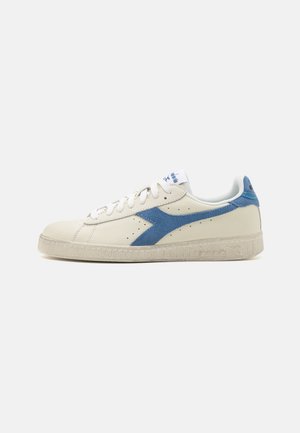 Sneakers in pelle crema con un accento in suede blu e suola in gomma texturizzata. Presentano lacci bianchi e un logo sulla linguetta.