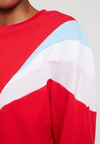 Sudadera roja con rayas superpuestas en azul claro, blanco y rojo adicional. Tejido suave con costuras visibles y cuello redondo.