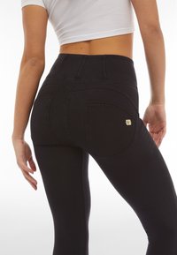 Nainen pukeutuneena korkeavyötäröisiin mustiin kapeisiin farkkuihin ja valkoiseen lyhyeen toppiin, seisomassa selin kameraan valkoista taustaa vasten.