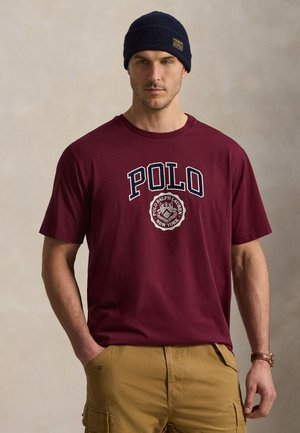 Mężczyzna w granatowej czapce typu beanie, bordowej koszulce Polo Ralph Lauren i jasnobrązowych spodniach cargo, stoi z jedną ręką w kieszeni na jednolitym tle.
