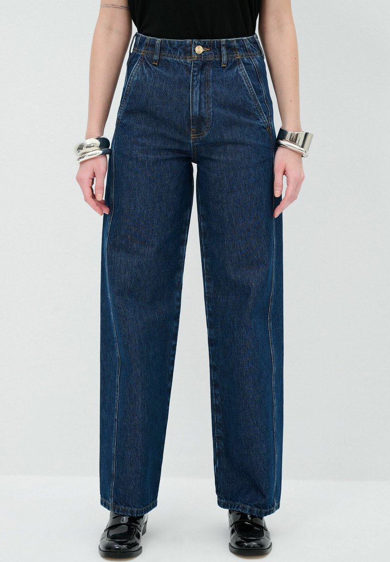 Džínové kalhoty s vysokým pasem a širokými nohavicemi v tmavě modré barvě. Mají oranžové švy, velké přední kapsy a zapínání na knoflík. Na nohou jsou lesklé černé boty.