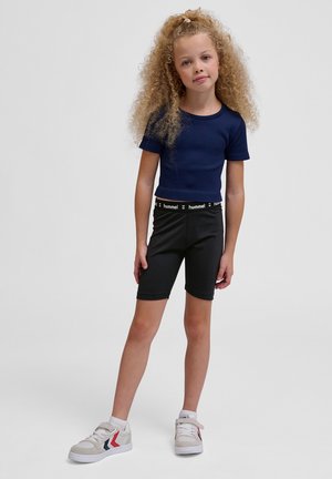 Marineblauw geribbeld shirt met korte mouwen, gecombineerd met zwarte skinny shorts met een logo op de tailleband. Model draagt witte sneakers met rode en blauwe accenten.