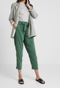 Pantaloni verdi larghi con vita regolabile; t-shirt grigia; blazer over-size a quadri bianco e nero; sandali neri con borchie.