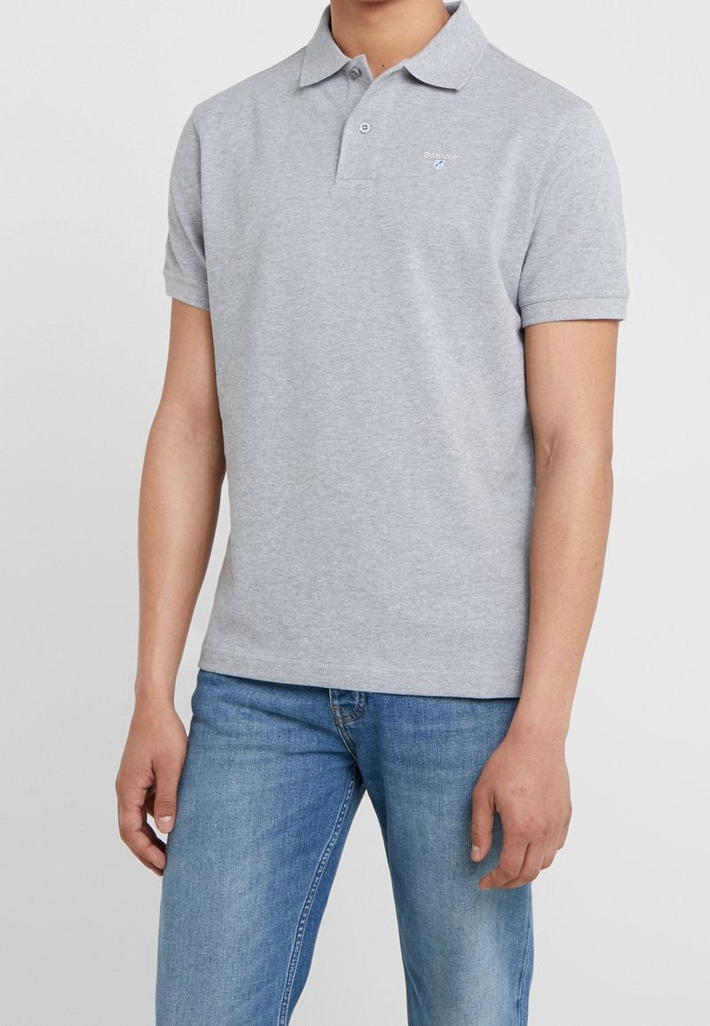 Homme portant un polo gris clair à manches courtes avec col, associé à un jean bleu en denim, debout devant un fond blanc uni.
