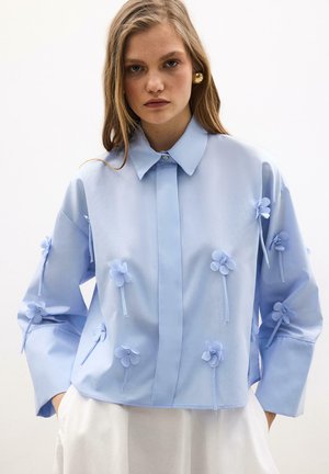 Mujer con una camisa de manga larga azul claro adornada con apliques florales en 3D, combinada con pantalones blancos sobre un fondo liso.