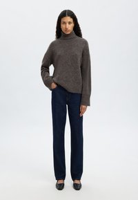 Szary sweter z wysokim kołnierzem, dziany, o luźnym kroju, noszony z ciemnoniebieskimi prostymi dżinsami i czarnymi płaskimi butami. Prosty, teksturowany wzór.