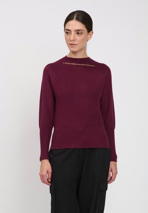 Kvinde med mørkt hår sat tilbage bærer en mørkerød ribstrikket sweater med en lille guldkædedetalje nær kraven og sorte bukser med lommer.
