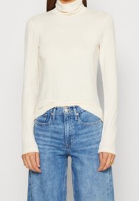 Crème turtleneck top van zacht materiaal, met lange mouwen en een strak ontwerp, gecombineerd met lichtblauwe wijde jeans.