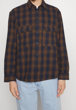 Uomo che indossa una camicia a quadri blu scuro e marrone con tasche sul petto, abbinata a jeans azzurri, in piedi contro uno sfondo bianco.