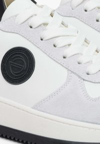 Białe sneakersy z szarymi zamszowymi akcentami, z okrągłą czarną łatą z logo, białymi sznurówkami i kontrastującą teksturą na cholewce. Czarna podeszwa.