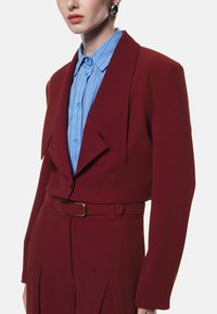 Blazer su misura color bordeaux con un colletto affilato, abbinato a una camicia a righe blu chiaro. Caratterizzato da una cintura ampia con accessori in oro e linee pulite.