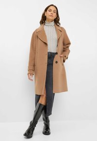 Tan uldblandingsfrakke med bred krave og sorte knapper. Båret over en mønstret turtleneck og en mørk denimnederdel; parret med skinnende sorte knæhøje støvler.