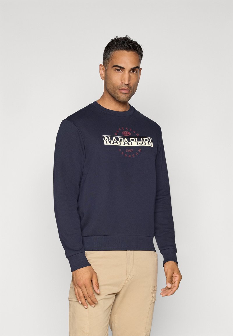 Homme portant un sweat-shirt bleu marine avec le texte "Napapijri" et un pantalon cargo beige, se tenant devant un fond clair uni.