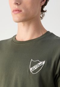 Olíva zöld póló rövid ujjakkal, kerek nyakkal, és hímzett fehér logóval a bal mellrészén, amely egy pajzs mintát ábrázol.