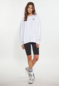 Felpa bianca oversized con testo ricamato nero, abbinata a pantaloni ciclistici neri. Il modello indossa sneaker bianche con dettagli neri e verdi.