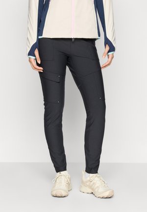 Svarte, tettsittende bukser med flere cargo-lommer, laget av stretch-stoff. Bært med en lett jakke og beige joggesko.