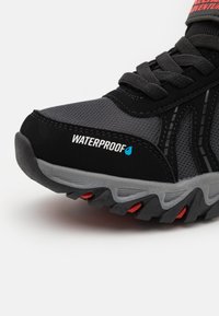Sneaker impermeabile nero e grigio con materiale in rete, superfici testurizzate e accenti rossi sulla suola. Presenta un design con lacci per una vestibilità sicura.
