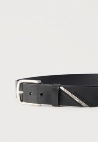 Melnas ādas josta ar sudraba metāla sprādzi, smalku tekstūru un sudraba zīmola birku ar šūšanas detaļu sānos.