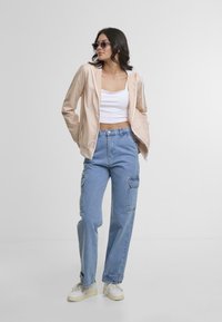 Ženska v svetlo rožnati zip-up jakni, beli kratki majici, modrih cargo kavbojkah, belih supergah in sončnih očalih, stoji ob navadni beli steni.