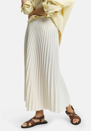 Person trägt einen cremefarbenen, plissierten Midirock, ein hellgelbes Langarmhemd mit hochgekrempelten Ärmeln und braune flache Sandalen.