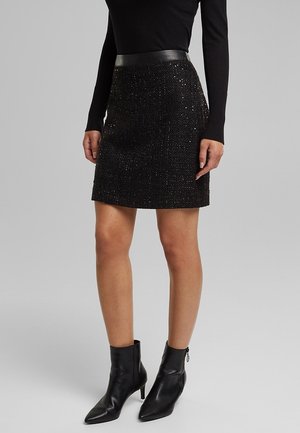 Frau trägt ein schwarzes langärmeliges Oberteil, einen strukturierten schwarzen Minirock mit Lederbund und schwarze spitze Stiefeletten mit Absatz.