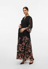 Long black maternity dress with floral print at the hem and sleeves. Features a tied waist, V-neck, and sheer long sleeves.