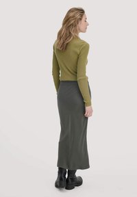 Frau mit langen blonden Haaren, die ein olivfarbenes, geripptes Langarmoberteil, einen dunkelgrünen Maxirock und schwarze, klobige Stiefel trägt, steht im Profil.