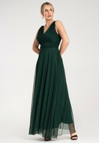 Długa sukienka w kolorze szmaragdowej zieleni, wykonana z lekkiego materiału, z plisowanym korpusem, dekoltem w kształcie litery V oraz zwiewnym dołem. Proste czarne sandały na cienkich obcasach.