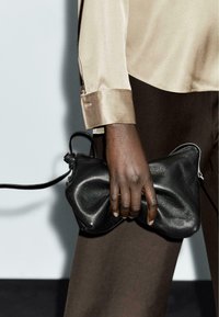 Clutch in pelle nera con una texture morbida e stropicciata e chiusura con zip. Il manico è sottile e si estende dal lato. Indossato da una mano.