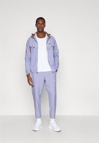 Άντρας που στέκεται φορώντας αθλητική φόρμα σε ανοιχτό μωβ χρώμα με λευκό t-shirt και λευκά αθλητικά παπούτσια, μπροστά σε ένα απλό ανοιχτό φόντο.