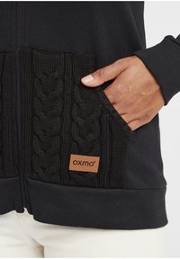 Čierna mikina na zips s pleteným káblovým vzorom na vreckách, koženou nášivkou loga, rebrovanými manžetami a jemnou textúrou. Detailný pohľad na ruku vo vrecku.