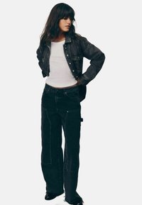Jachetă din denim închisa la culoare, purtată cu un top alb ajustat și blugi negri cu picioare largi, având buzunare frontale și detalii de cusătură. Modelul stă în profil.
