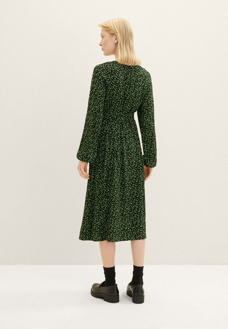 Grünes Blumen-Midi-Kleid mit langen Ärmeln, taillierter Passform und ausgestelltem Saum, kombiniert mit schwarzen Socken und klobigen Schuhen. Glatte Stoffstruktur.