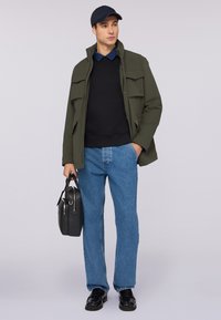 Chaqueta llamativa verde con bolsillos grandes, suéter negro, vaqueros azules, zapatos negros y un bolso negro estructurado, combinado con una gorra azul marino.