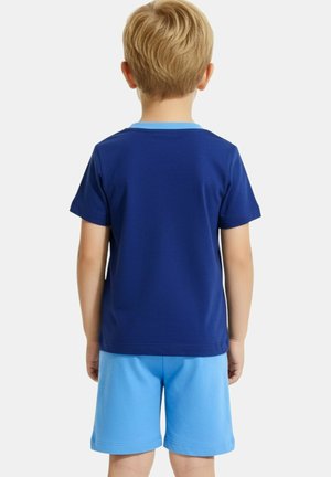 Un enfant blond est vu de dos, portant une chemise à manches courtes bleu marine et un short bleu clair sur un fond blanc.