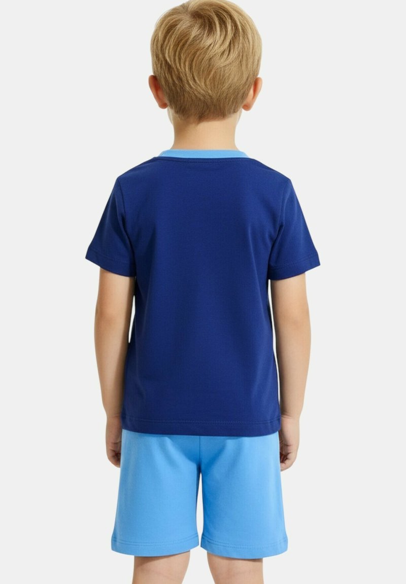Un enfant blond est vu de dos, portant une chemise à manches courtes bleu marine et un short bleu clair sur un fond blanc.