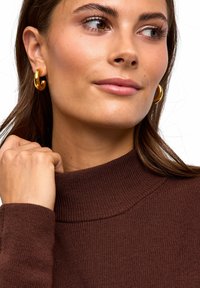 Brauner gerippter Rollkragenpullover mit hohem Ausschnitt, getragen mit großen goldenen Creolen. Die Haut ist glatt und betont das natürliche Make-up.