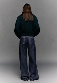 Debela tamnozelena pletena majica širokih rukava, uparena s tamnoplavim trapericama širokih nogavica visokog struka, s džepovima na stražnjoj strani i kontrastnim šavovima.