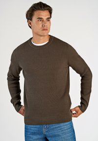 Suéter marrón de canalé con cuello redondo, acabado texturizado, mangas ajustadas y corte recto. Combinado con jeans de denim azul.