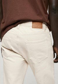 Jeans in denim beige con una texture liscia, dotati di due tasche posteriori e un'etichetta in pelle marrone sulla vita.