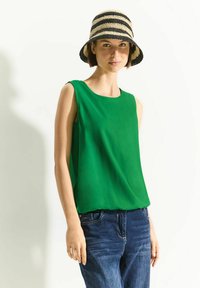 Top sans manches vert en tissu lisse, avec un col rond, associé à un jean en denim bleu foncé et un chapeau de paille à rayures.